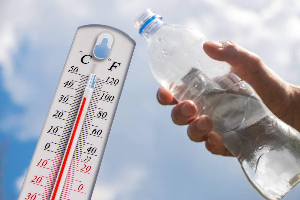 Hand hält Trinkflasche und im Hintergrund ein Thermometer welches eine hohe Temperatur anzeigt